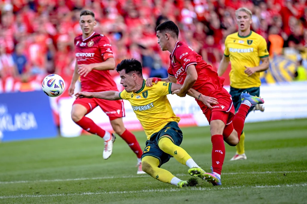 Dynamiczna akcja podczas meczu piłkarskiego, zawodnik w żółtej koszulce przewraca się na murawie walcząc o piłkę z przeciwnikiem w czerwonym stroju, w tle widoczni kolejni piłkarze oraz kibice na trybunach.