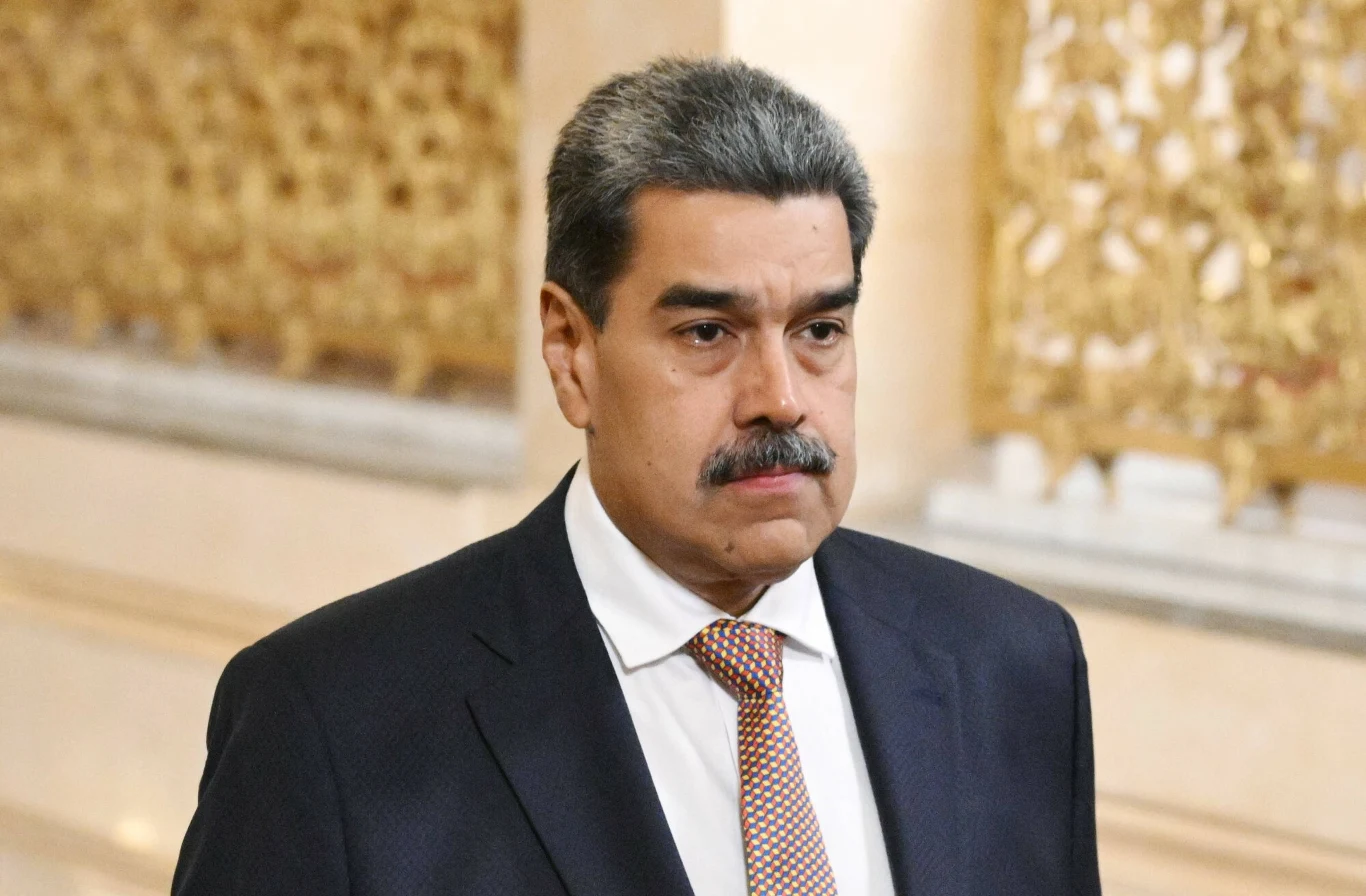 Nicolasa Maduro Mężczyzna o poważnym wyrazie twarzy, ubrany w ciemny garnitur, białą koszulę oraz krawat w drobną kratkę, stoi w eleganckim wnętrzu z ozdobnymi złotymi elementami w tle.