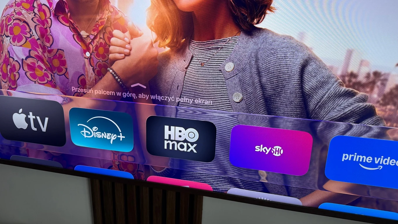 Apple TV za pół ceny na Black Friday. Promocja na aż sześć miesięcy. Ekran telewizora wyświetlający aplikacje serwisów streamingowych takich jak Apple TV, Disney+, HBO Max, Sky oraz Prime Video na tle fragmentu wizerunku dwóch osób.