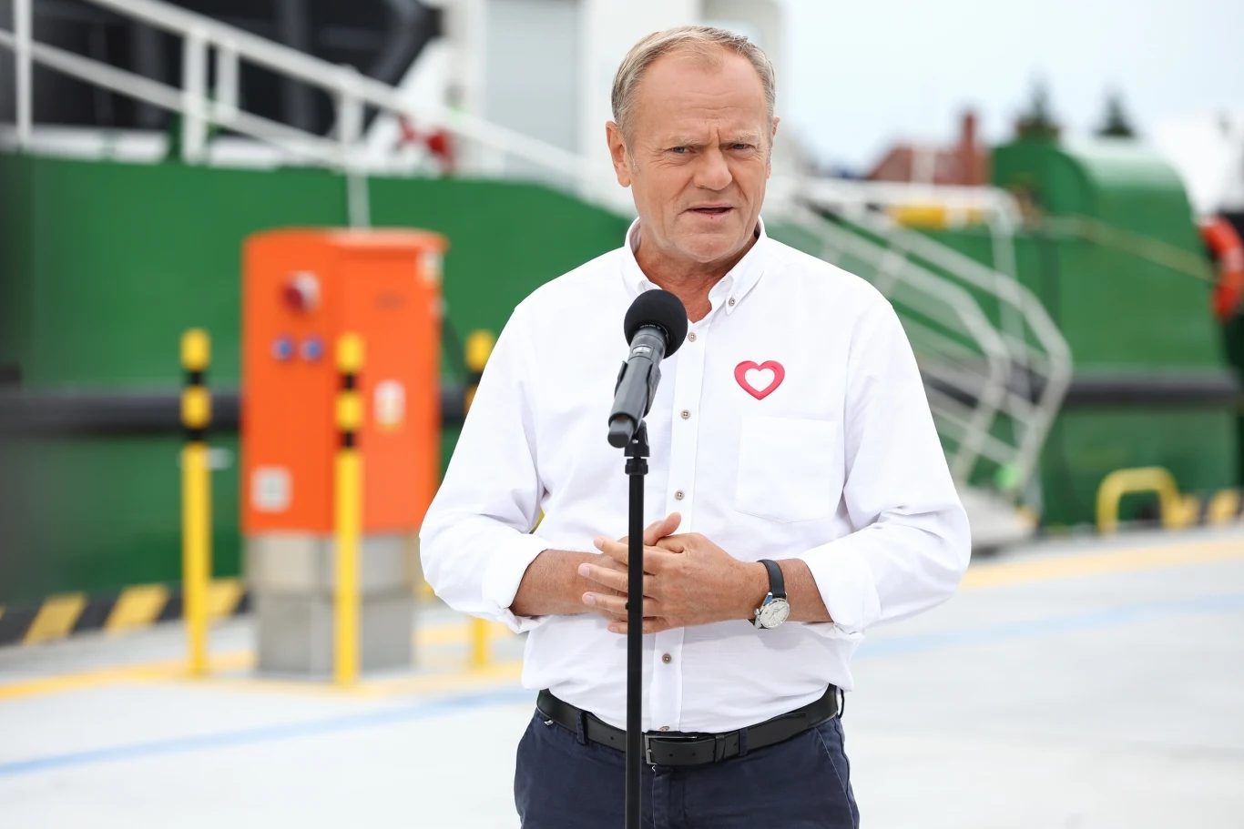 Donald Tusk reaguje na burzę wokół KPO Mężczyzna w białej koszuli z przypiętym sercem przemawia do mikrofonu na tle infrastruktury przemysłowej i pomarańczowego urządzenia technicznego.