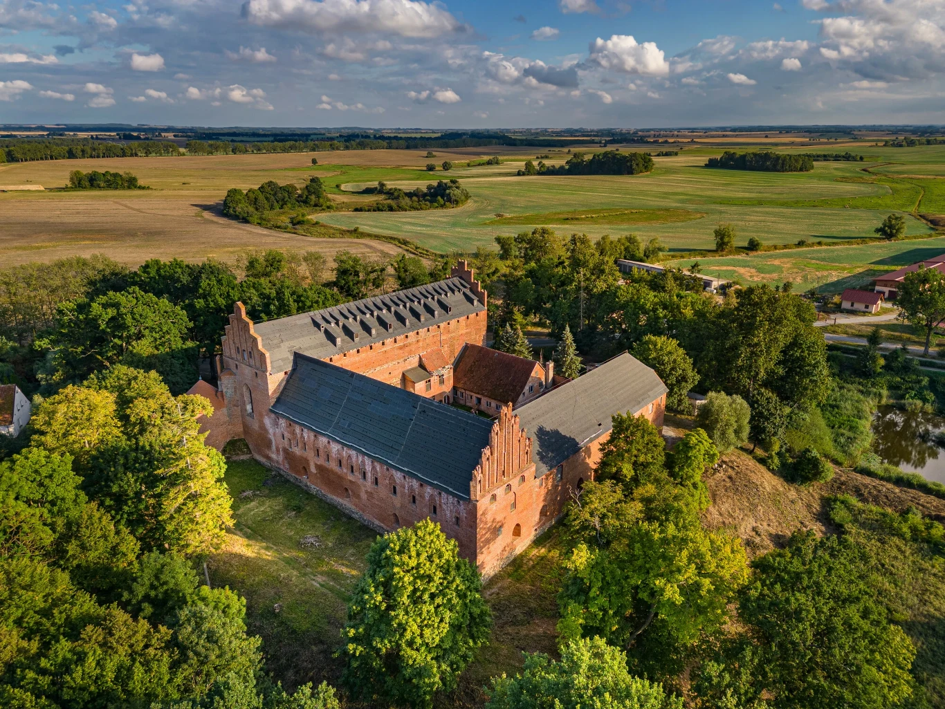 Zamek w Barcianach wciąż na sprzedaż. Cena rośnie Gotycki zamek otoczony zielenią drzew, położony na niewielkim wzniesieniu, z widokiem na rozległe pola uprawne pod błękitnym niebem z nielicznymi chmurami.