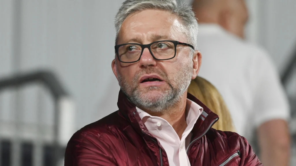 Jerzy Brzęczek Mężczyzna w okularach i bordowej kurtce, z siwą brodą i krótkimi włosami, patrzy w bok na tle rozmytego wnętrza z innymi osobami w tle.