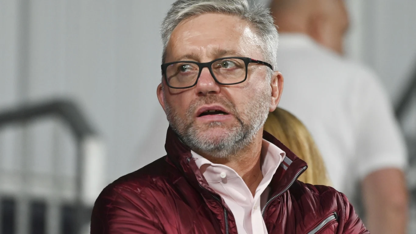 Jerzy Brzęczek Mężczyzna w okularach i bordowej kurtce, z siwą brodą i krótkimi włosami, patrzy w bok na tle rozmytego wnętrza z innymi osobami w tle.