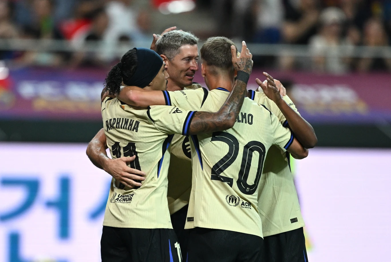 Lewandowski świętujący z kolegami z drużyny kolejne trafienie Grupa piłkarzy w jasnych koszulkach obejmuje się wzajemnie na boisku podczas celebracji po zdobytym golu, widoczne emocje i radość, w tle rozmyta publiczność na trybunach.