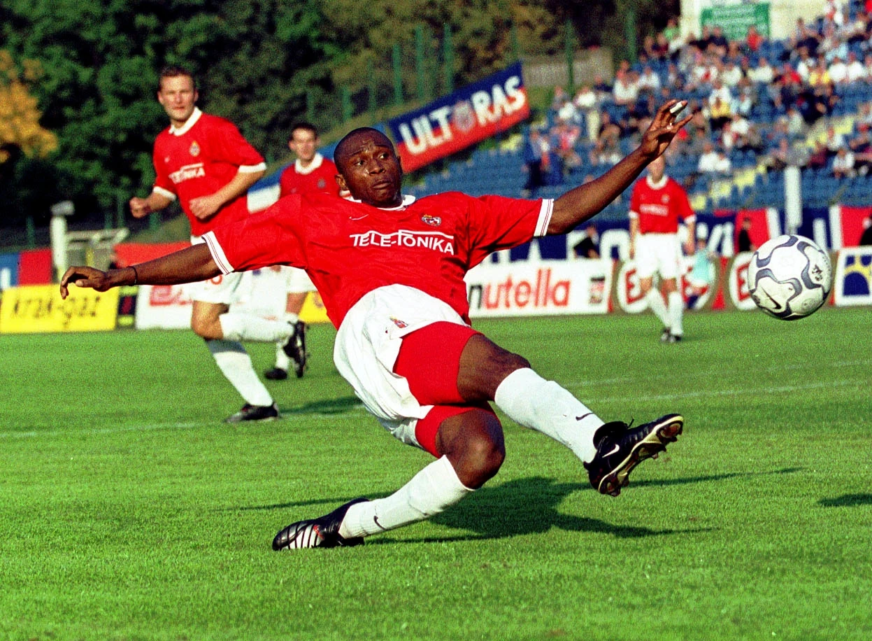 Zawodnik w czerwono-białym stroju piłkarskim wykonuje dynamiczny wślizg na murawie, zbliżając się do piłki podczas meczu, w tle widoczni są inni gracze i trybuny pełne kibiców.