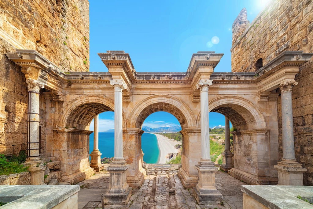 Brama Hadriana znajduje się w samym centrum kurortu Antalya Krągłe łuki i kolumny starożytnych ruin z widokiem na nadmorską panoramę, jasne niebo oraz lazurowa woda w tle.