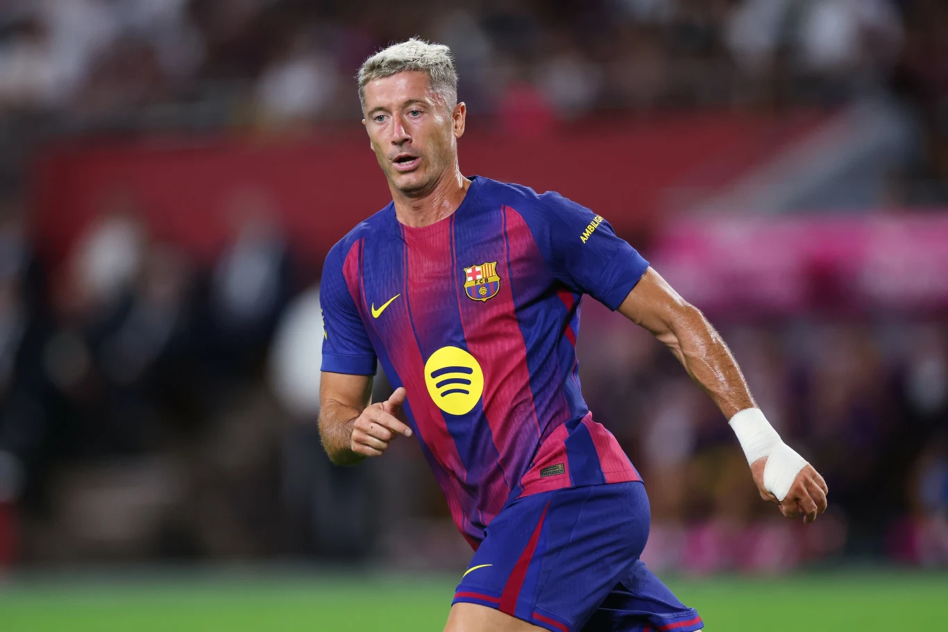 Robert Lewandowski Piłkarz w stroju FC Barcelony z widocznym logiem sponsora na koszulce, w ruchu na boisku podczas meczu, dynamiczna sylwetka sportowca o wyróżniającym się jasnym kolorze włosów, w tle rozmyta publiczność stadionu.