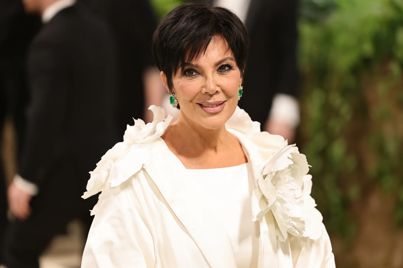 Kris Jenner nie wygląda na swój wiek, podobnie jak jej matka Kobieta z krótkimi, ciemnymi włosami uśmiecha się delikatnie, ubrana w elegancką, białą kreację z ozdobnymi kwiatowymi elementami na ramionach, ma zielone kolczyki, w tle rozmazane sylwetki i naturalna zieleń.