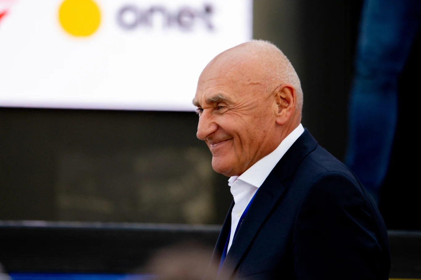 Starszy, łysy mężczyzna w eleganckim garniturze, uśmiechnięty, zwrócony bokiem, stoi na tle nowoczesnego wnętrza z widocznym logo Onet w tle.