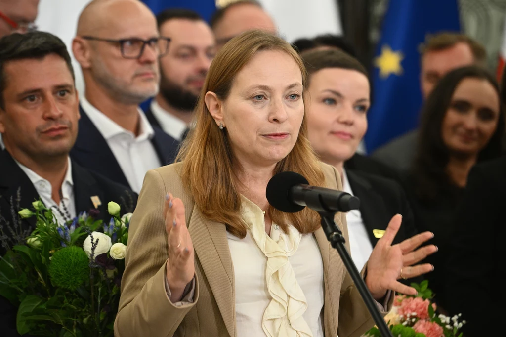Kobieta przemawia do mikrofonu gestykulując rękami, w tle znajduje się grupa osób oraz kwiaty, a także widoczne są flagi, w tym flaga Unii Europejskiej.