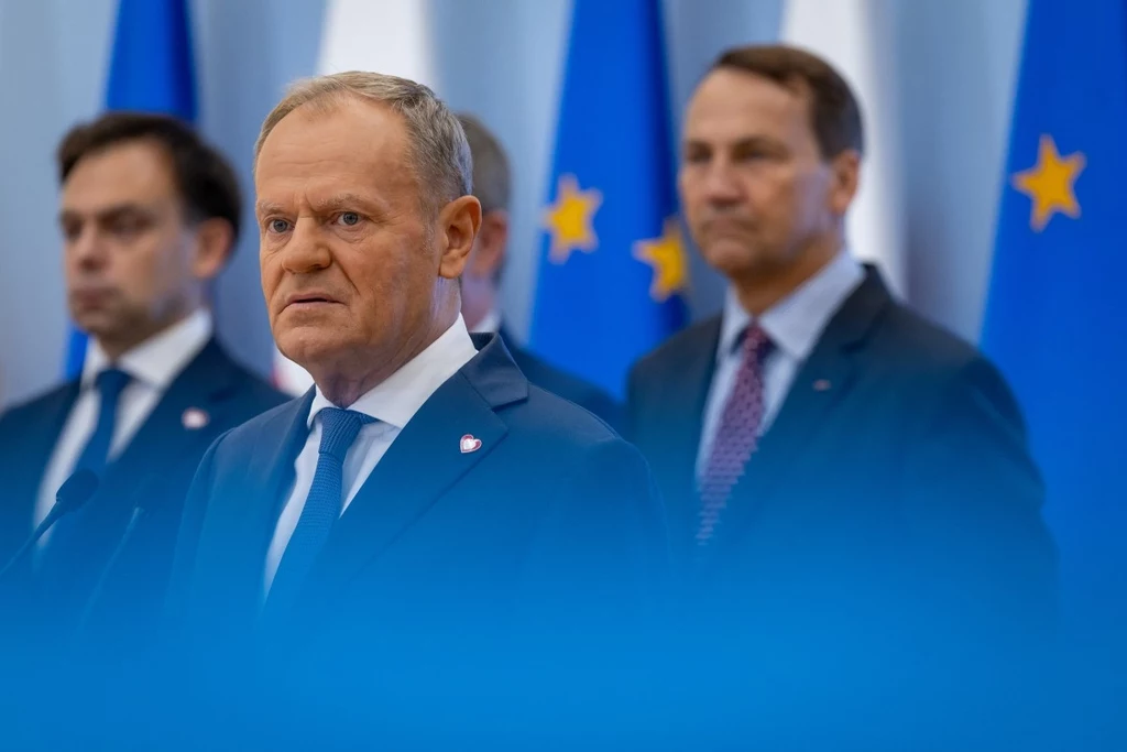 Na pierwszym planie: premier Polski Donald Tusk Trzech mężczyzn w garniturach na tle flag Unii Europejskiej i Polski, na pierwszym planie skupiony mężczyzna z siwiejącymi włosami, pozostali w tle lekko nieostro, całość utrzymana w oficjalnej atmosferze.