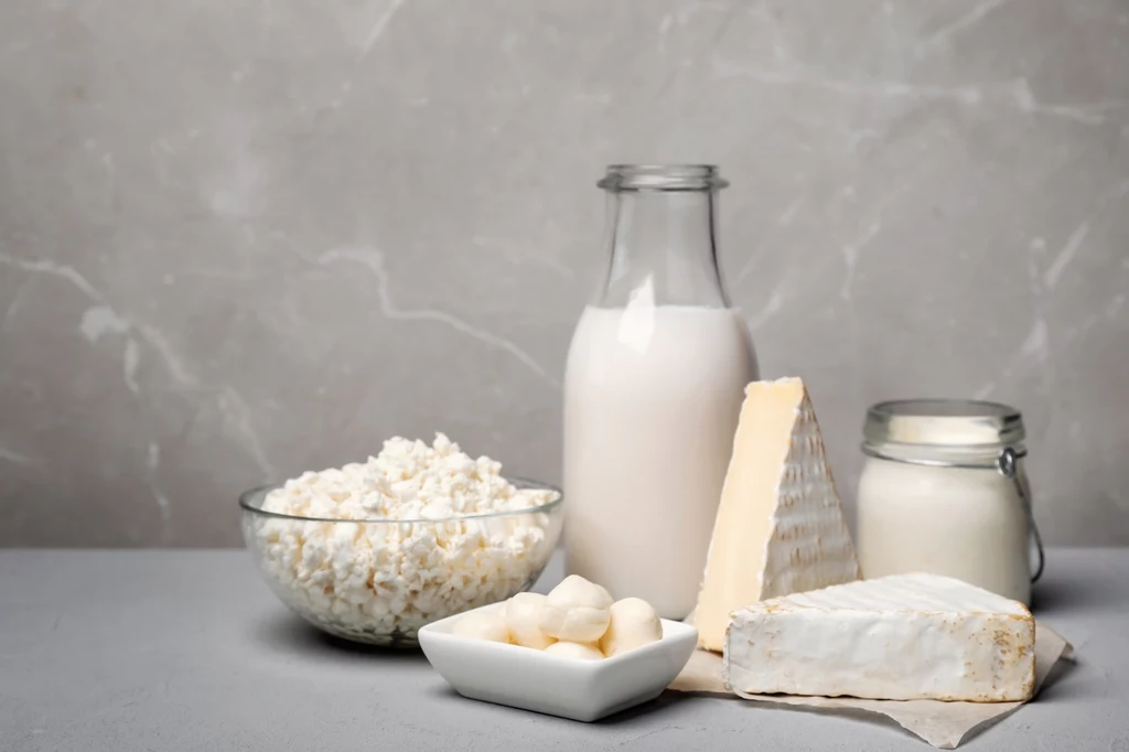 Szklana butelka mleka w otoczeniu różnych produktów mlecznych, takich jak twaróg w misce, ser camembert, twardy ser dojrzewający oraz jogurt w słoiku ustawione na jasnym tle.