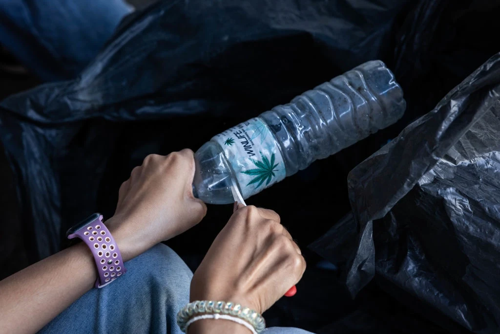Czy trzeba usuwać etykiety z plastikowych butelek? Ręce osoby trzymającej plastikową butelkę z naklejonym rysunkiem liścia marihuany oraz napisem, jedna z rąk wycina fragment butelki przy pomocy nożyczek, w tle ciemne torby foliowe.