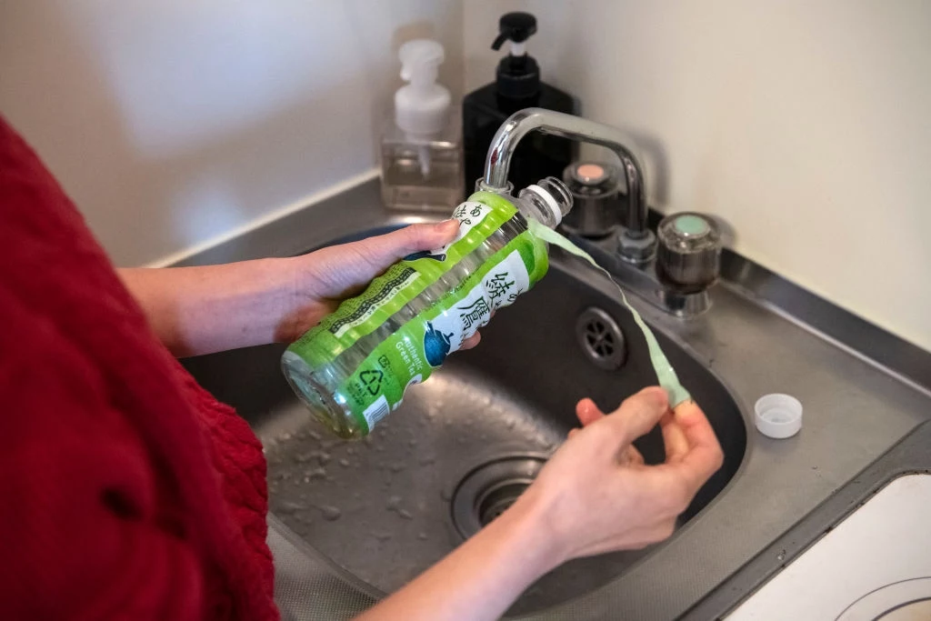 Ręce trzymają plastikową butelkę po płynie do zmywania, do której nalewana jest woda z kranu, na tle zlewu kuchennego z widocznymi innymi pojemnikami z płynami.
