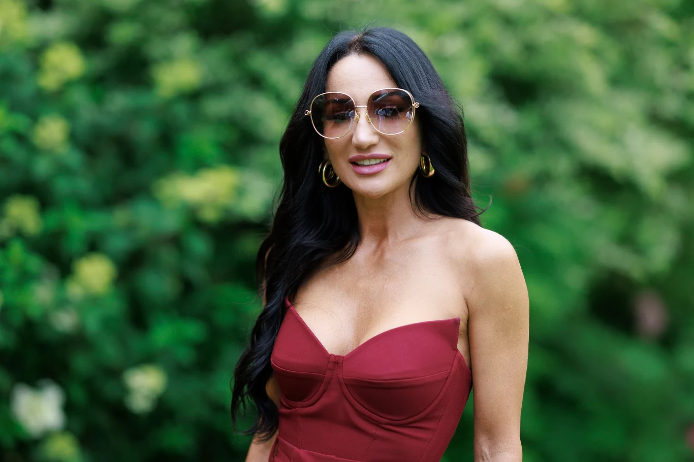 Justyna Steczkowska Kobieta z długimi czarnymi włosami w bordowej sukience, dużych okularach przeciwsłonecznych oraz złotych kolczykach stoi na tle zieleni.