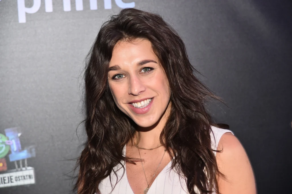 Joanna Jędrzejczyk Młoda kobieta z długimi, ciemnobrązowymi włosami uśmiecha się, ubrana w jasną sukienkę, stoi na tle ciemnej ścianki z nieczytelnym logo.