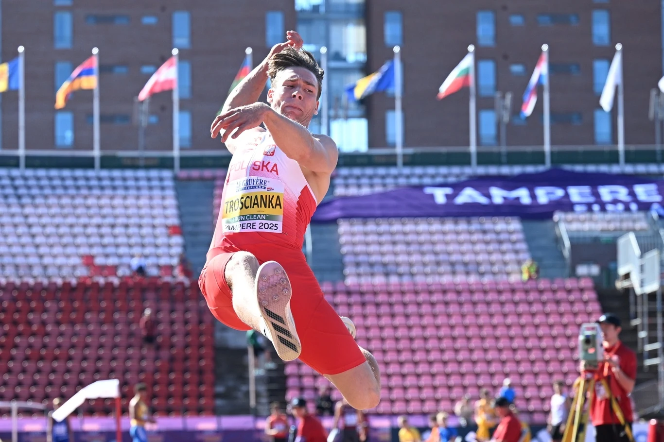 Lekkoatleta w biało-czerwonym stroju wykonuje skok w dal na stadionie lekkoatletycznym, w tle widoczne trybuny, flagi różnych państw oraz napisy związane z wydarzeniem sportowym.