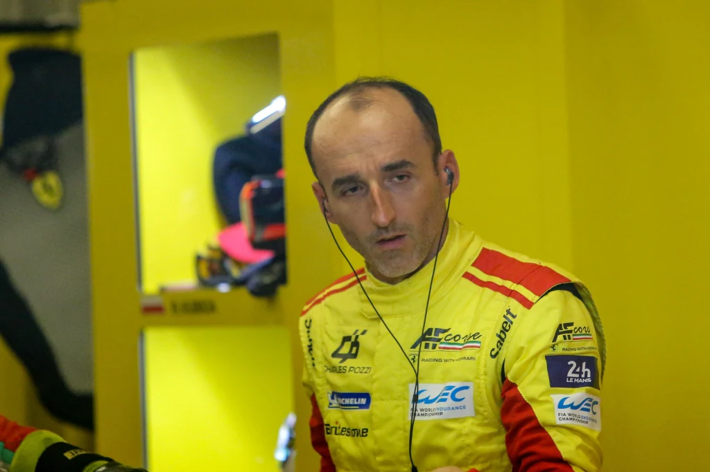 Robert Kubica Kierowca wyścigowy ubrany w żółty kombinezon zespołu wyścigowego, z zestawem słuchawkowym na uszach, stoi w boksie wyścigowym na tle żółtych szafek; na kombinezonie widoczne są logotypy sponsorów.