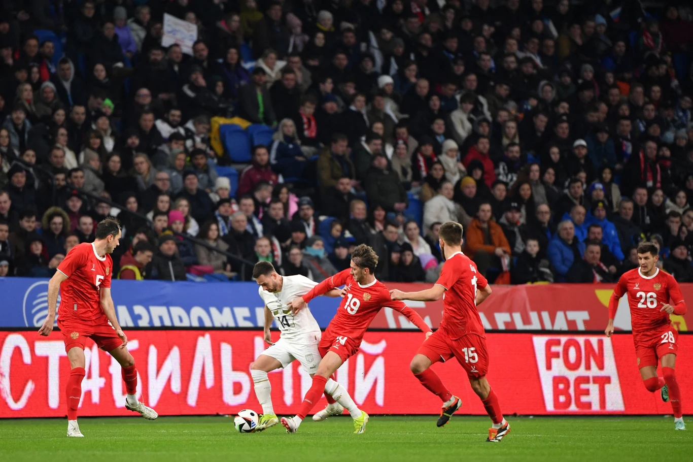 Piłkarz w białej koszulce drybluje z piłką na tle obrońców w czerwonych strojach, wokół niego czterech zawodników próbuje przejąć piłkę, w tle widoczny tłum kibiców na trybunach.