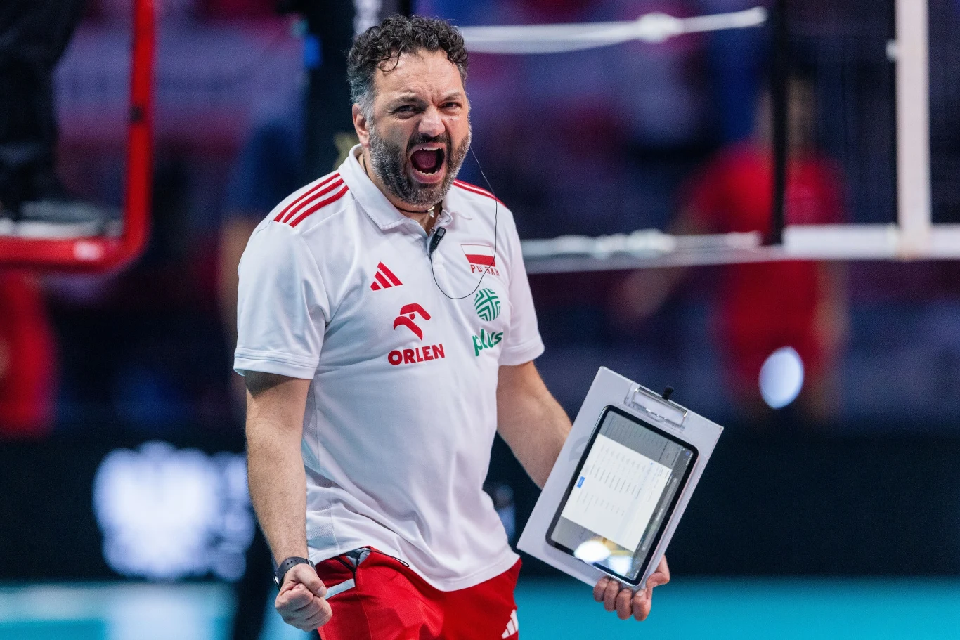 Trener w białej koszulce i czerwonych spodenkach wyraża silne emocje, trzymając w ręku tablicę z notatkami na tle hali sportowej. Jego postawa sugeruje zaangażowanie i energię związane z wydarzeniem sportowym.