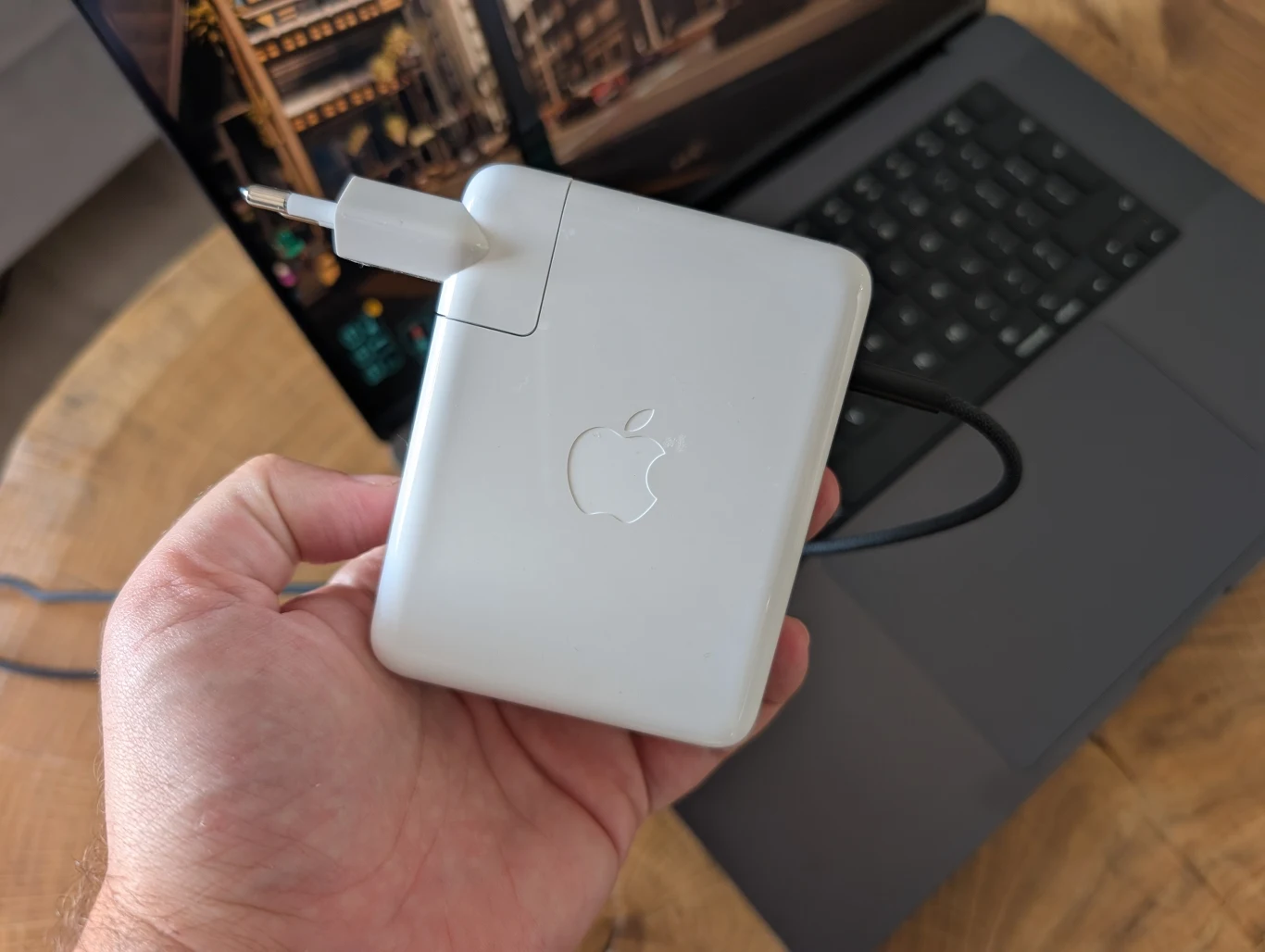 MacBook Pro 16" z M4 Pro Ręka trzyma białą ładowarkę do laptopa marki Apple, z widocznym kablem i charakterystycznym logo, w tle częściowo widoczny laptop oraz blat stołu.