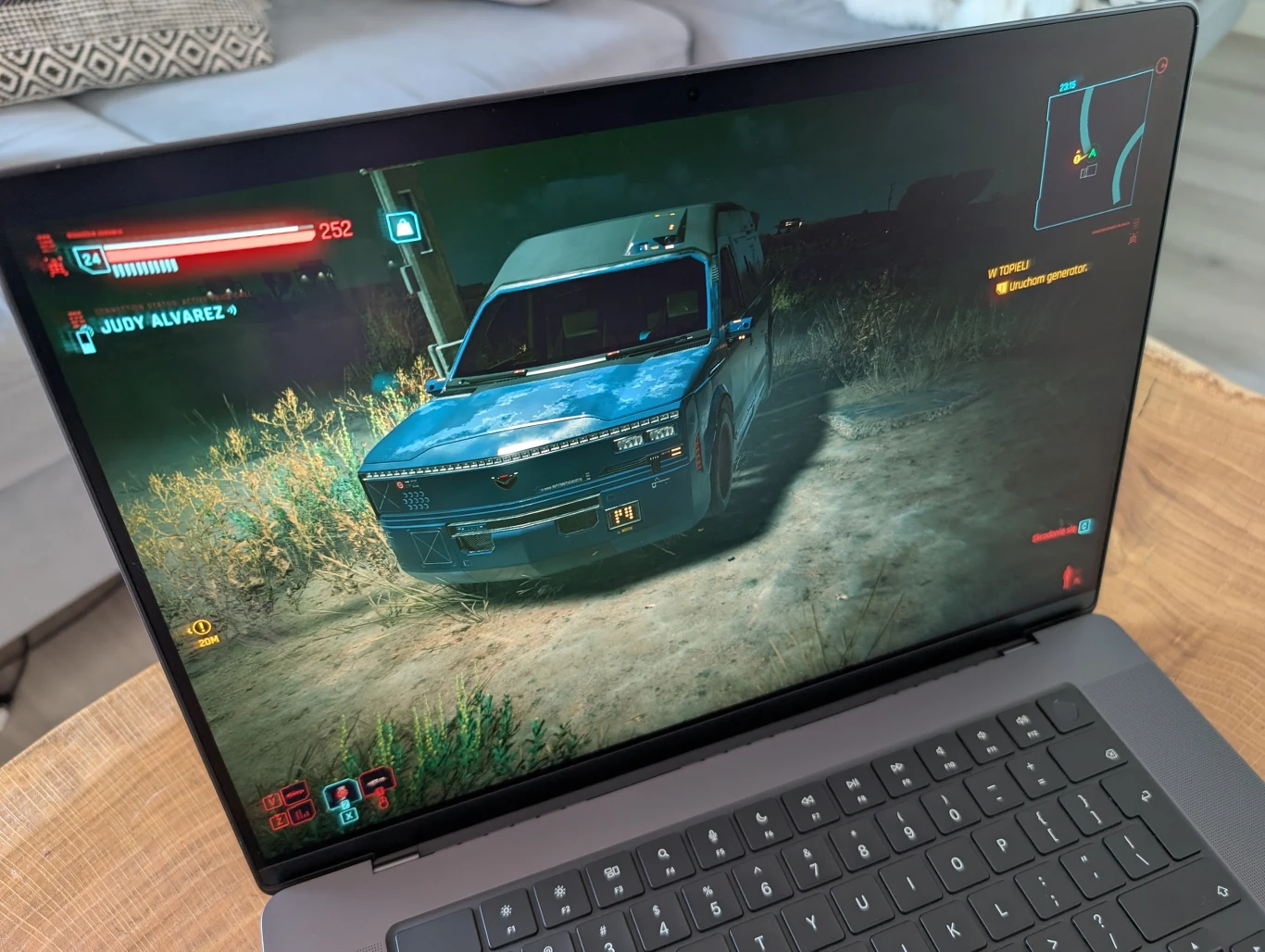 MacBook Pro 16" z M4 Pro Ekran laptopa ukazujący rozgrywkę w grze komputerowej, na której znajduje się niebieski pojazd zaparkowany na tle roślinności, elementy interfejsu gry i mapa widoczne w lewym i prawym górnym rogu ekranu.