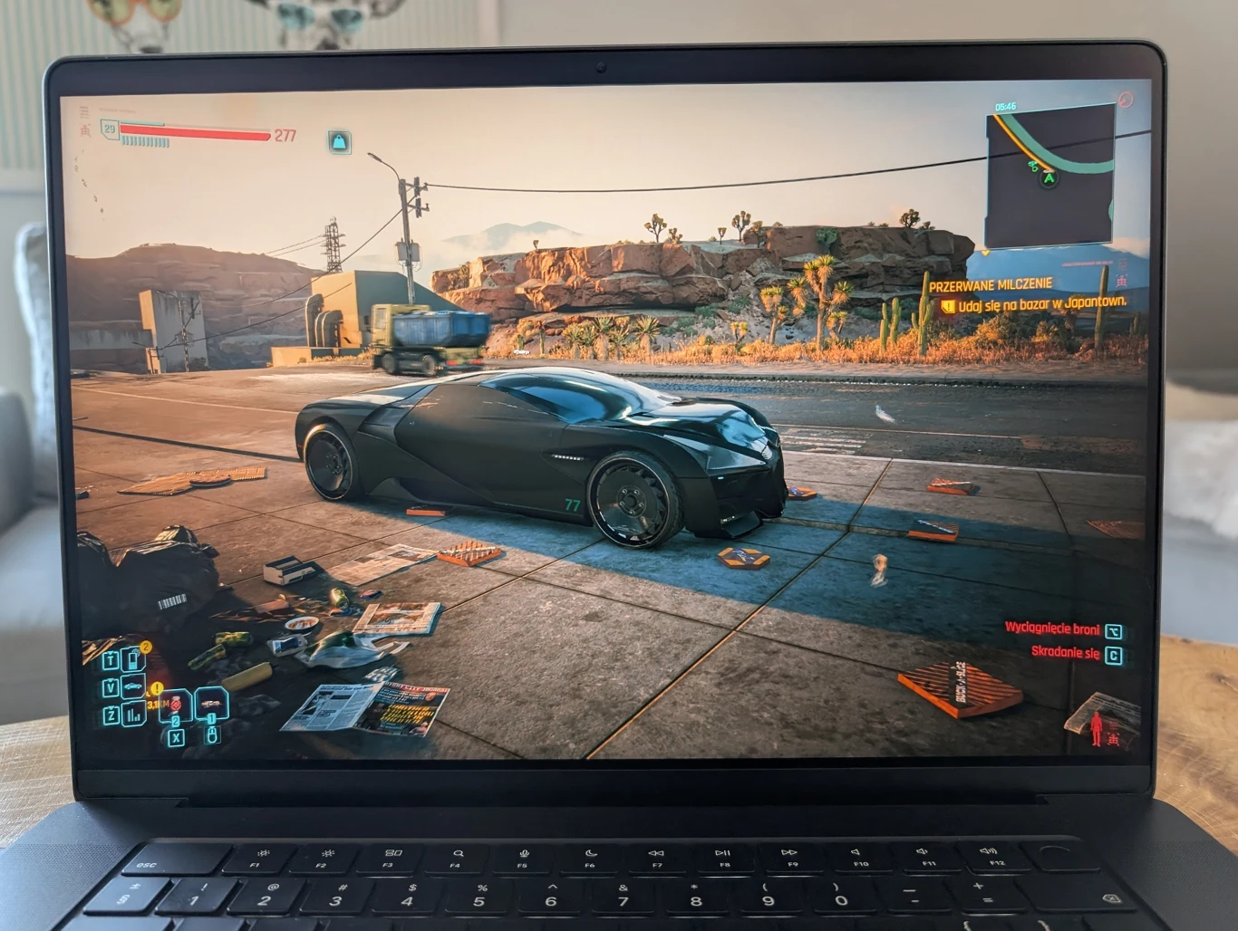 MacBook Pro 16" z M4 Pro Ekran laptopa wyświetlający scenę z gry komputerowej, gdzie na ulicy stoi futurystyczny czarny samochód sportowy, otoczony rozrzuconymi przedmiotami, w tle widoczny krajobraz pustynny oraz fragmenty asfaltowej drogi.