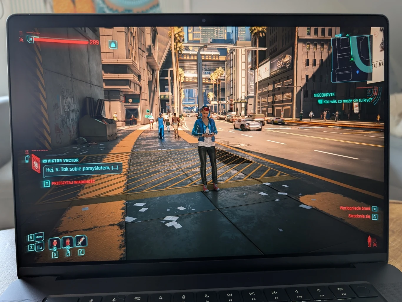 MacBook Pro 16" z M4 Pro Widok ekranu laptopa przedstawiającego grę komputerową Cyberpunk 2077, na którym widać postać stojącą na ulicy futurystycznego miasta otoczonego wysokimi budynkami, interfejsem gry oraz elementami cyberpunkowej architektury.