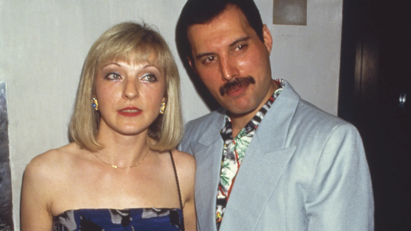 Mary Austin i Freddie Mercury Para eleganckich osób pozuje razem, kobieta w sukience na ramiączkach i mężczyzna w jasnej marynarce z kwiecistą koszulą.