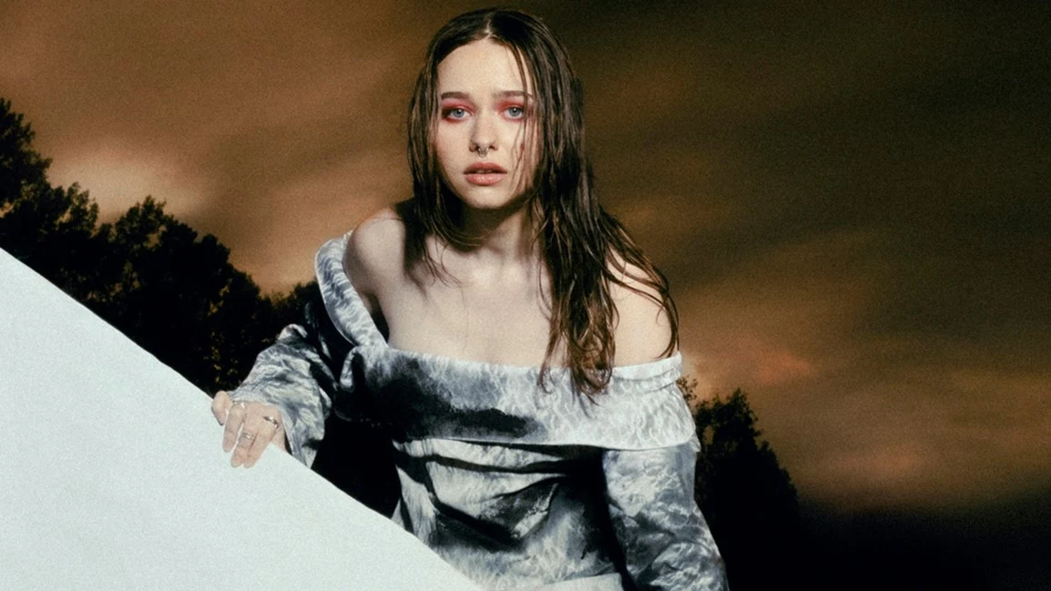 Livka Kobieta z mokrymi włosami i rozmazanym makijażem oczu, ubrana w srebrną, obszerą suknię z odsłoniętymi ramionami, stoi na tle ciemnego, burzowego nieba i sylwetek drzew.