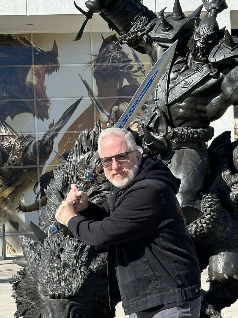 Rod Fergusson Mężczyzna w okularach przeciwsłonecznych pozuje z mieczem przed dużą rzeźbą postaci fantasy przedstawiającą wojownika w zbroi i bestię, tło stanowi mural z motywami fantasy.
