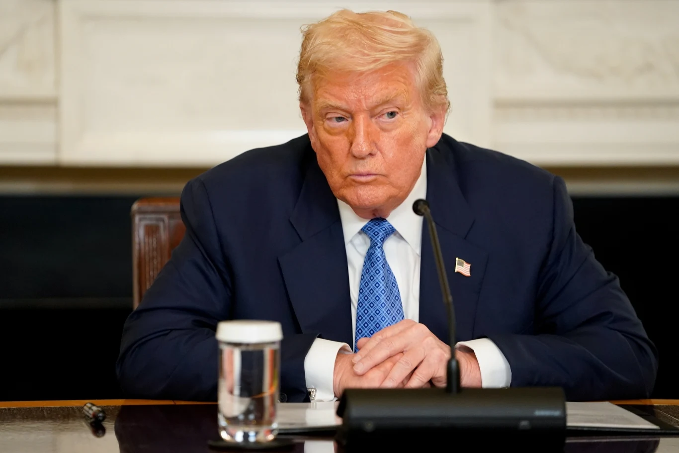 Prezydent USA Donald Trump Mężczyzna w garniturze siedzący przy stole, z poważnym wyrazem twarzy, przed nim znajduje się szklanka wody oraz mikrofon, w tle fragment jasnej ściany.