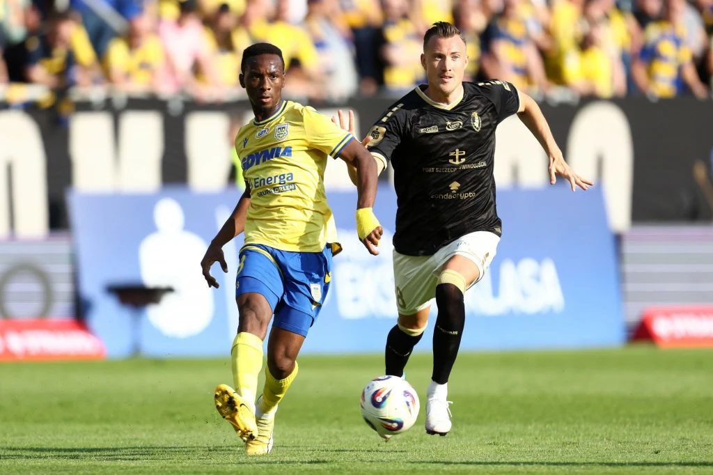 Arka Gdynia - Pogoń Szczecin Dwóch piłkarzy podczas dynamicznej rozgrywki na boisku, jeden z nich w żółto-niebieskim stroju biegnie obok zawodnika w czarnym stroju, na pierwszym planie piłka, w tle widownia oraz banery reklamowe.