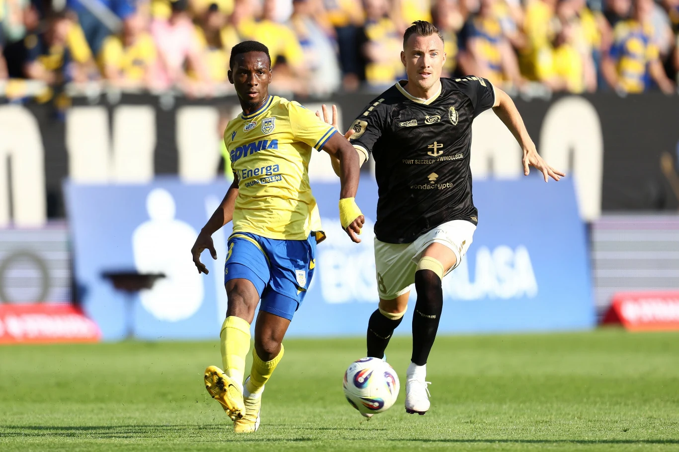 Dwóch piłkarzy podczas dynamicznej rozgrywki na boisku, jeden z nich w żółto-niebieskim stroju biegnie obok zawodnika w czarnym stroju, na pierwszym planie piłka, w tle widownia oraz banery reklamowe.