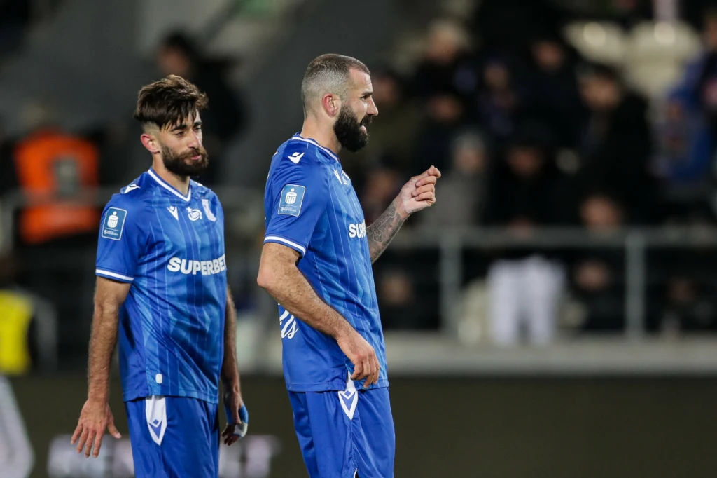 Dwóch piłkarzy w niebieskich strojach sportowych z napisem 'SUPEREBET' stoi na murawie, jeden z nich odwrócony bokiem, drugi patrzy prosto, w tle zamazana publiczność i trybuny stadionu.