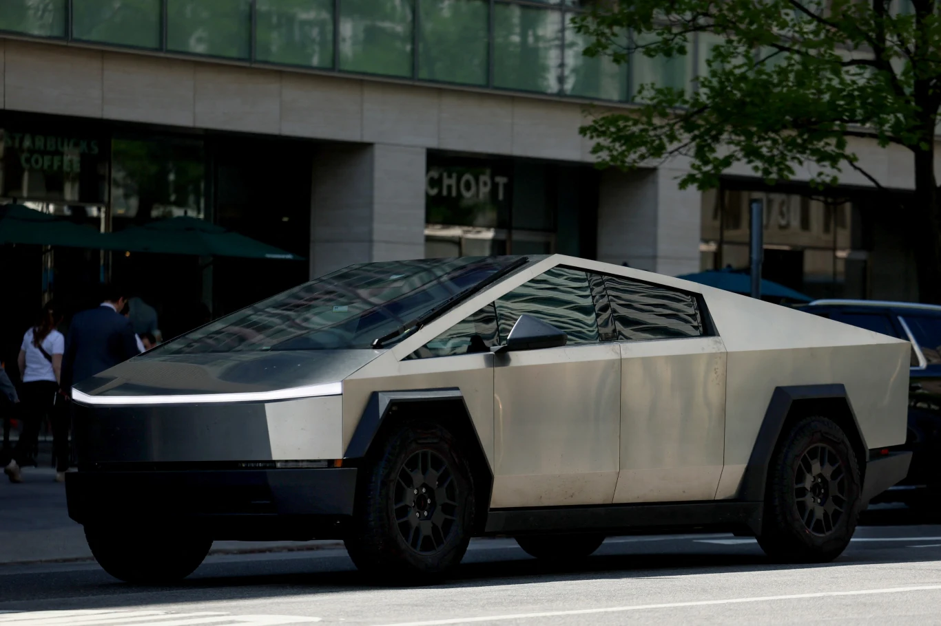Samochód Tesla Cybertruck Nowoczesny samochód elektryczny o futurystycznym, kanciastym kształcie zaparkowany przy ulicy w miejskim otoczeniu, w tle przeszklony budynek i drzewa rzucające cień.