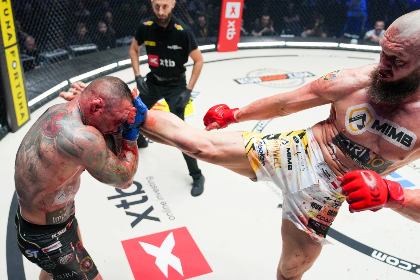 Jeden zawodnik MMA otrzymuje kopnięcie w głowę od przeciwnika podczas walki w oktagonie, obaj są mocno umięśnieni i pokryci tatuażami, na ciele widoczne ślady po ciosach, tło stanowi siatka klatki i obserwujący sędziowie.