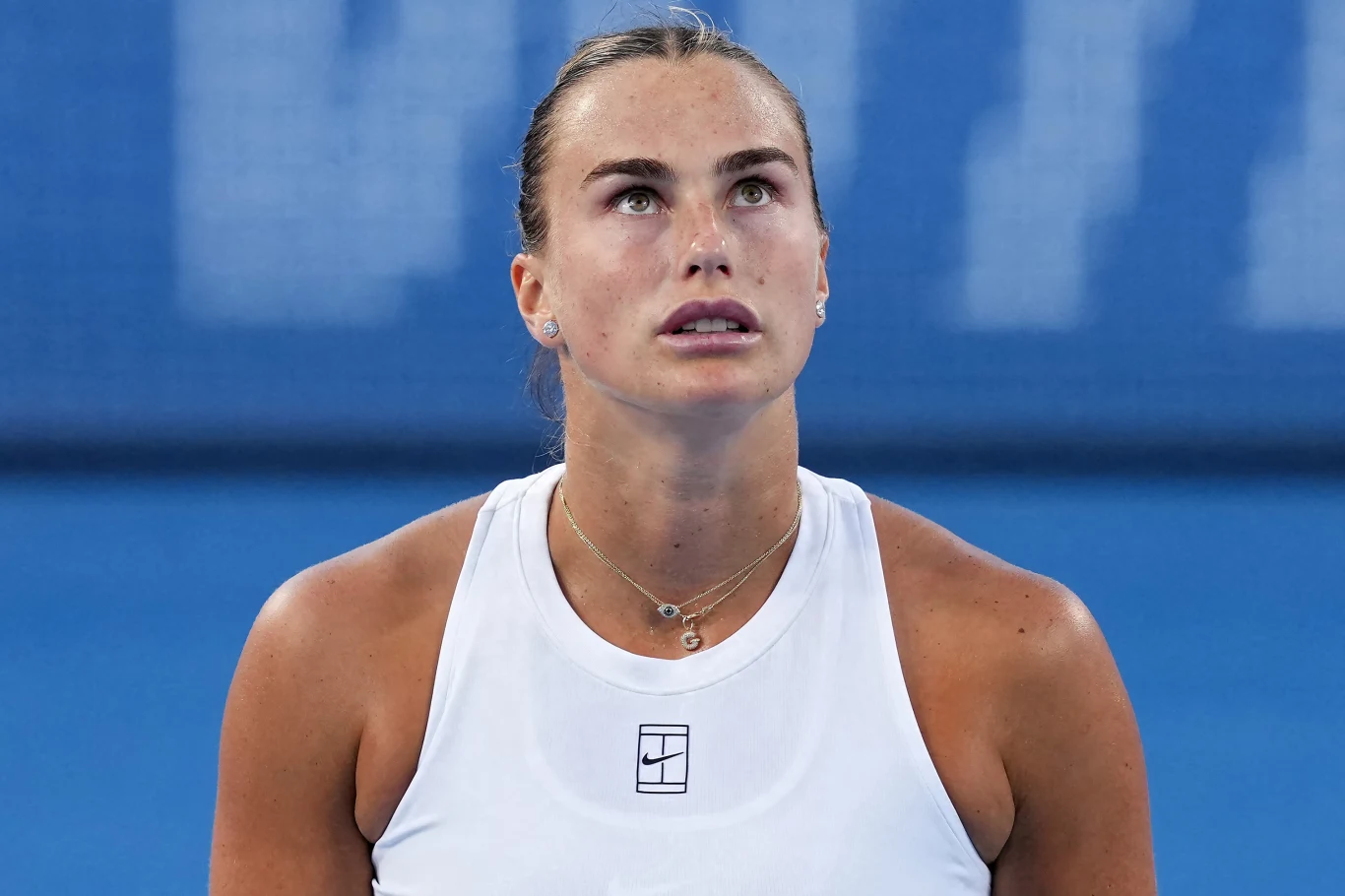 Aryna Sabalenka Kobieta w sportowym stroju, patrząca w górę z poważnym wyrazem twarzy, na tle rozmytego niebieskiego obiektu sportowego.