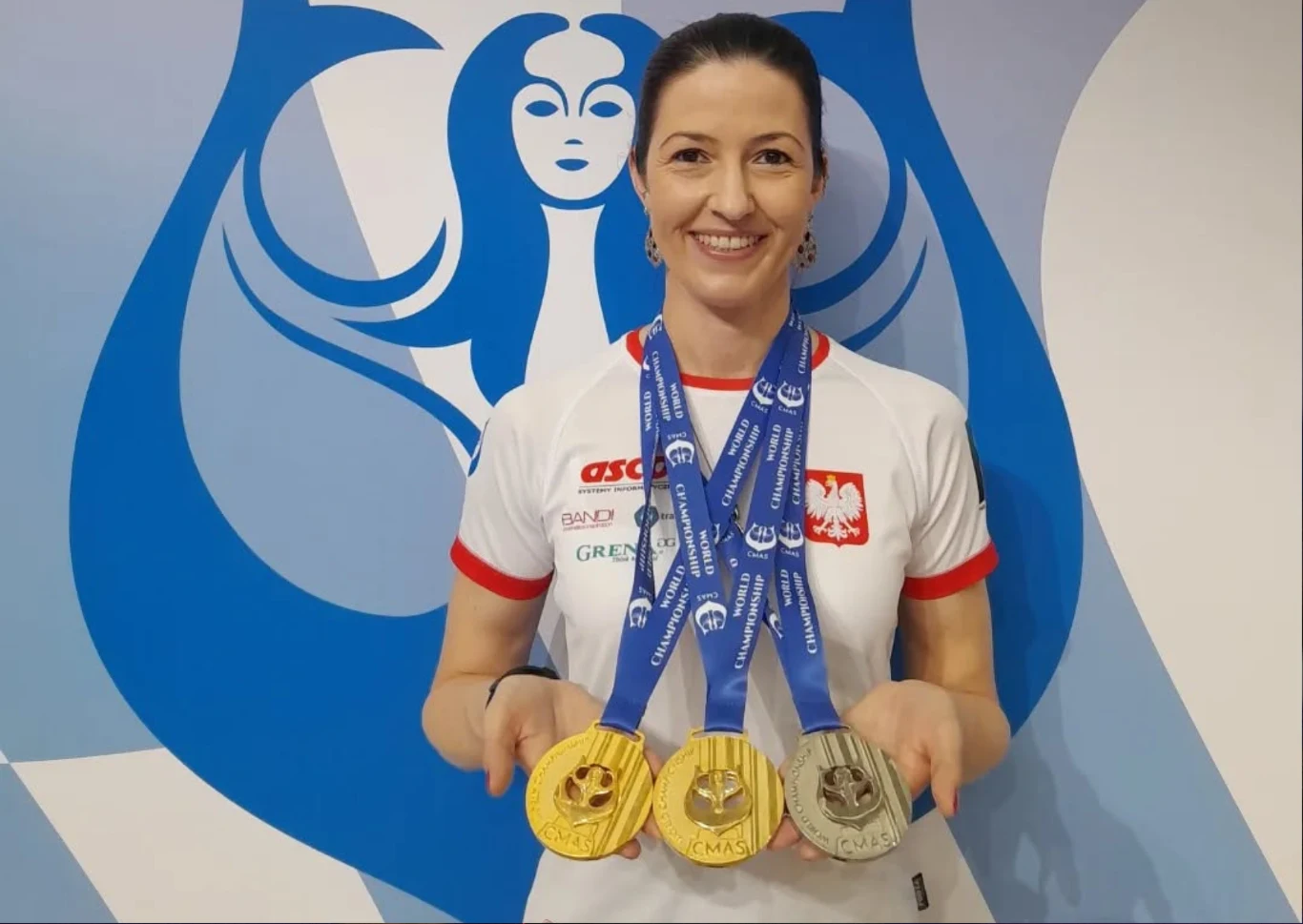 Julia Kozerska z medalami za zwycięstwa podczas CMAS World Championship 2025 Kobieta ubrana w biały sportowy T-shirt z godłem Polski, trzymająca trzy medale, w tle niebiesko-białe graficzne logo przedstawiające stylizowaną postać kobiecą.