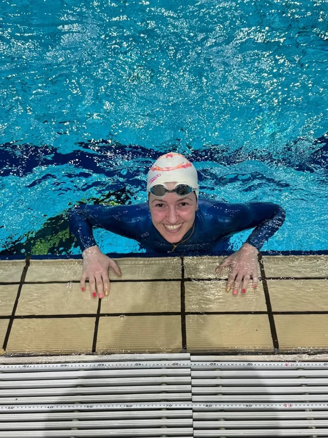 Julia Kozerska Uśmiechnięta kobieta w stroju pływackim, czepku i okularach do pływania trzyma się brzegu basenu, w tle woda o jasnoniebieskim odcieniu