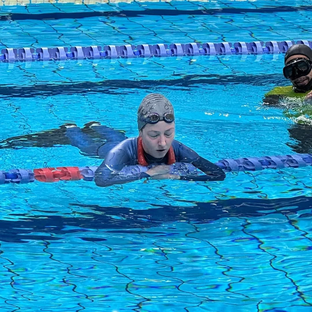 Julia Kozerska Osoba w czepku i okularach pływackich odpoczywa przy krawędzi toru pływackiego w basenie, obok w wodzie znajduje się druga osoba w masce.