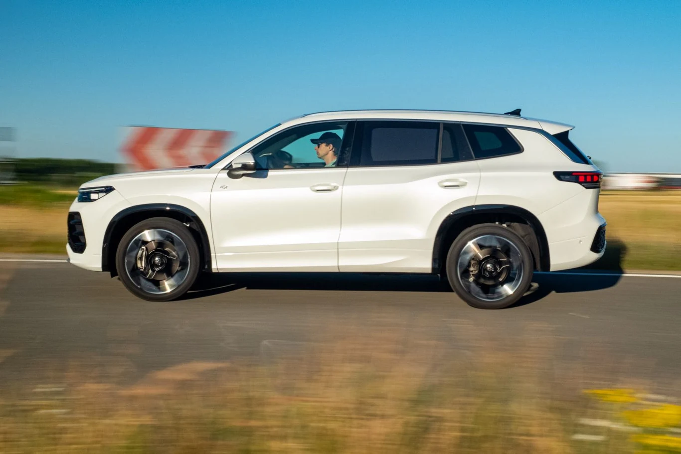 Volkswagen Tayron jest o ponad 25 cm dłuższy od Tiguana - różnice w wyglądzie obu modeli najmocniej widać z boku Biały SUV jadący drogą, z widocznym kierowcą za kierownicą, w tle rozmazany krajobraz i znak drogowy świadczący o ruchu, auto w dynamicznym ujęciu bocznym.