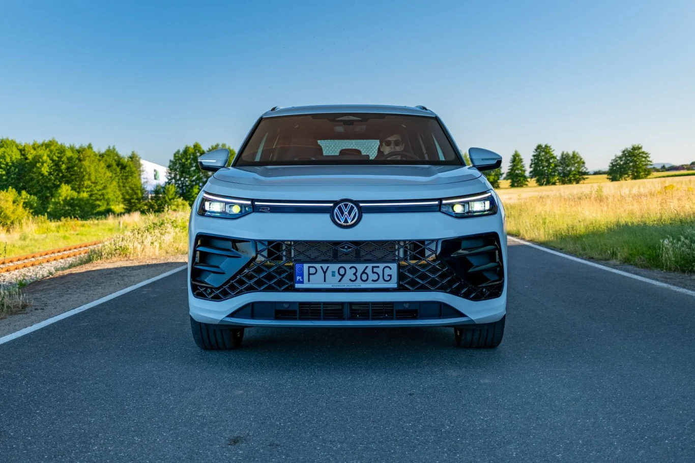 Tayron, jak większość współczesnych Volkswagenów, ma w przedniej części nadwozia podświetlany pas łączący reflektory Nowoczesny samochód marki Volkswagen ustawiony na asfalcie, w otoczeniu zielonych pól i błękitnego nieba, z widoczną tablicą rejestracyjną oraz reflektorami LED.