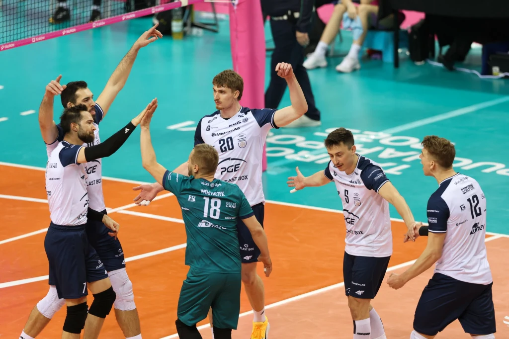 Grupa siatkarzy w sportowych strojach świętuje zdobyty punkt na boisku do siatkówki, unosząc ręce i okazując radość; parkiet w odcieniach pomarańczowego i zielonego, siatka oraz trybuny w tle.