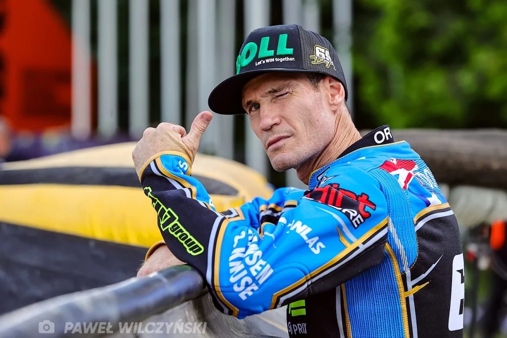 Jason Doyle Mężczyzna w stroju wyścigowym z bejsbolówką na głowie, stojący przy ogrodzeniu i pokazujący gest kciuka w górę oraz mrugający jednym okiem, w tle widoczne barierki i elementy infrastruktury sportowej.
