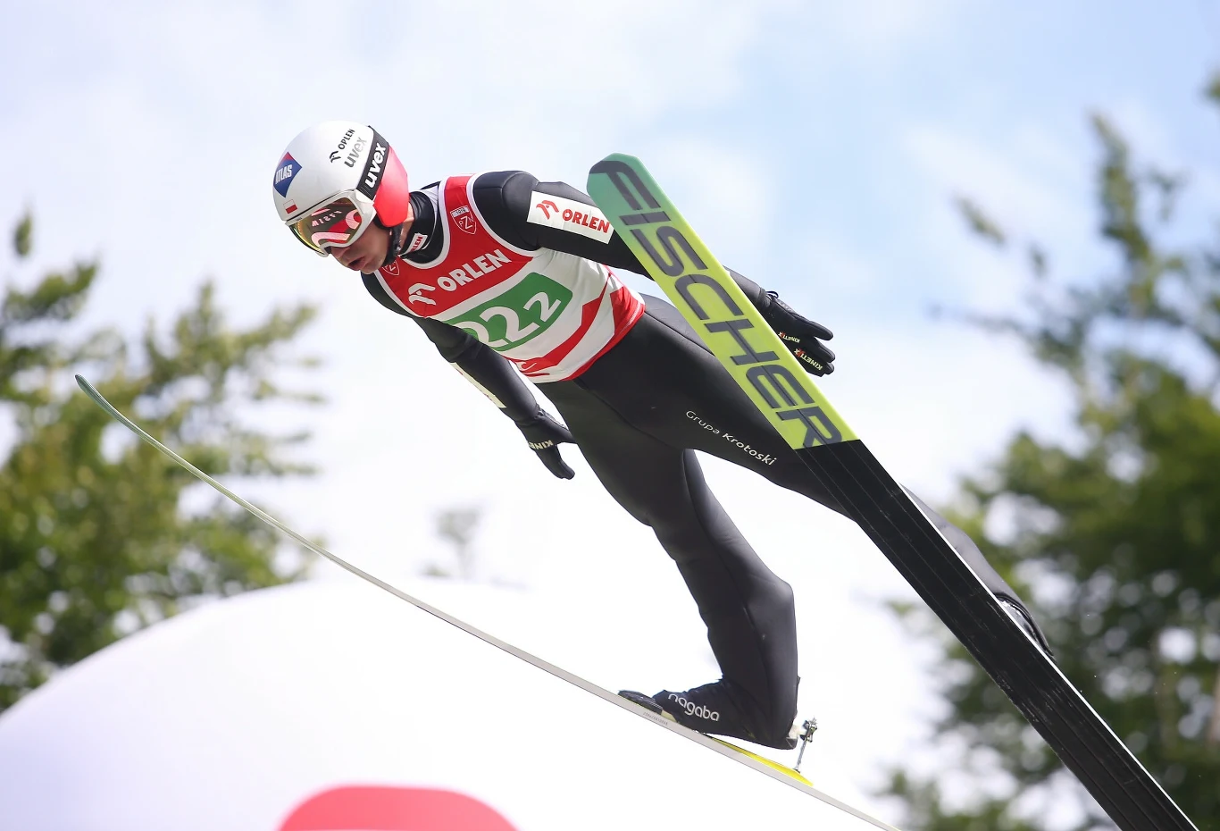 Skoczek narciarski w locie na tle błękitnego nieba, ubrany w kombinezon sportowy z numerem startowym i kaskiem ochronnym, narty uniesione równolegle, w tle rozmyte drzewa i elementy skoczni.