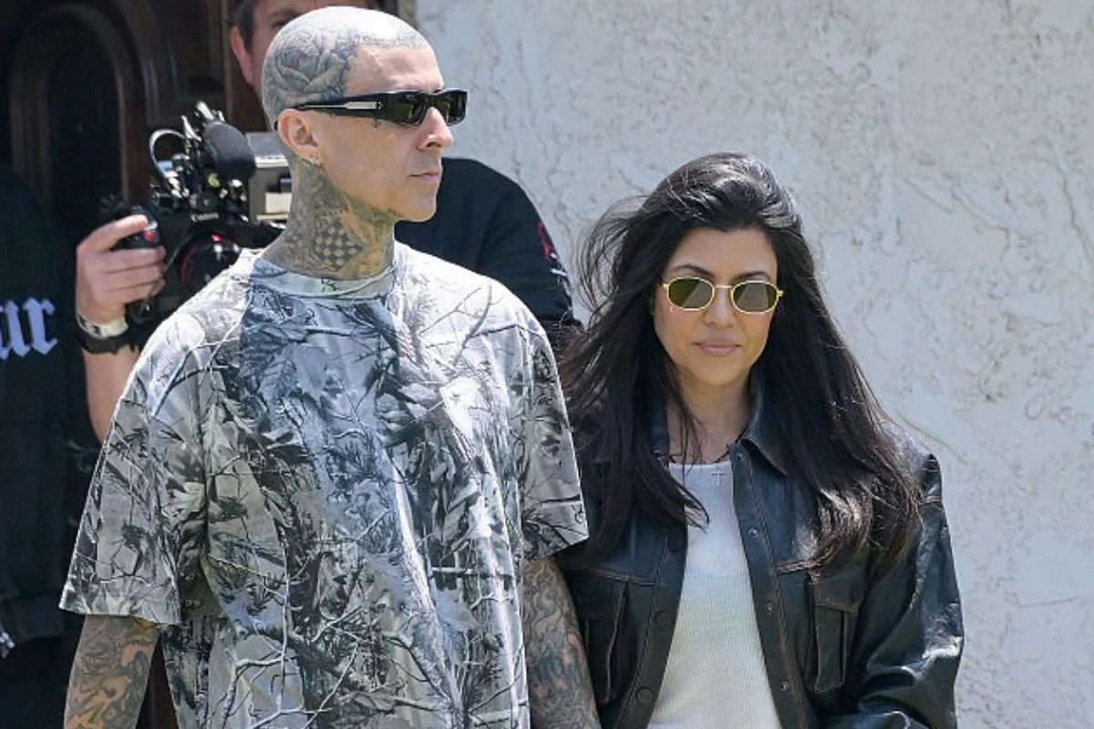 Kourtney Kardashian z mężem Travisem Barkerem Mężczyzna z tatuażami na głowie i szyi w ciemnych okularach oraz kobieta o długich ciemnych włosach w skórzanej kurtce i również w okularach przeciwsłonecznych stoją obok siebie na tle jasnej ściany.