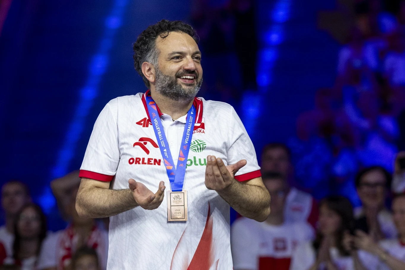 Stefano Lavarini Mężczyzna w białej koszulce z logotypami sponsorów, z medalem na niebieskiej szarfie na szyi, uśmiecha się i gestykuluje rękami, w tle kibice w biało-czerwonych barwach.