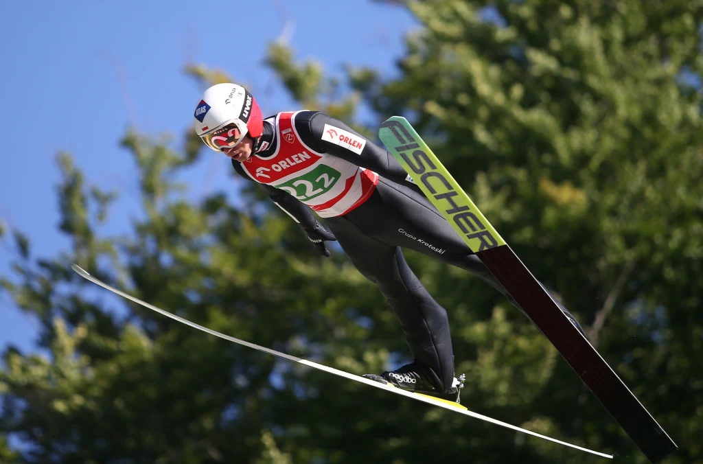 Kamil Stoch Skoczek narciarski w czerwono-czarnej kombinacji w locie na tle jasnoniebieskiego nieba i zielonych drzew, dynamiczna pozycja wskazuje na środek skoku podczas zawodów.