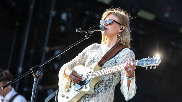 Kobieta w okularach przeciwsłonecznych gra na elektrycznej gitarze i śpiewa do mikrofonu podczas występu na scenie na świeżym powietrzu, ubrana w białą koronkową sukienkę, za nią widoczny drugi muzyk.